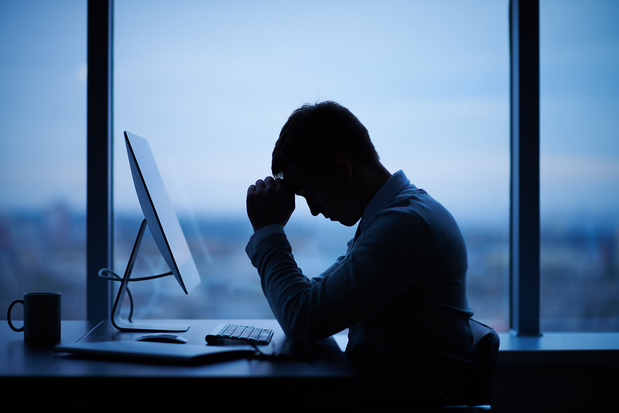 a man having problems while sat down to his chair with a computer & a mug on his desk